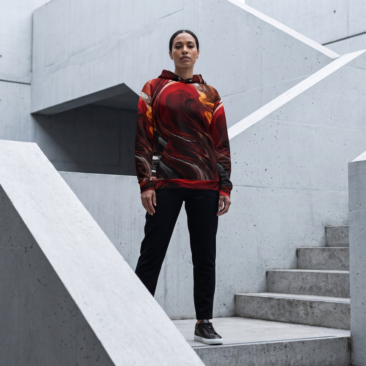 A woman stands on concrete stairs wearing a long-sleeve hoodie with a red, orange, and black abstract flame-like print, paired with black pants and dark sneakers.