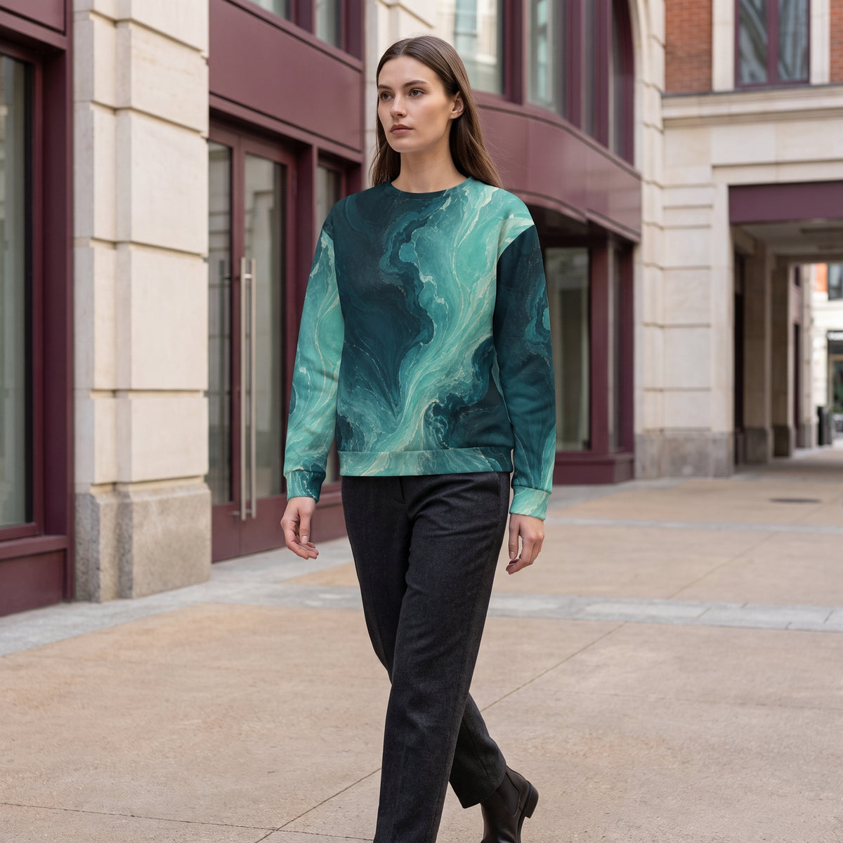 A woman walking outdoors on a city sidewalk wearing a long-sleeve crewneck sweatshirt with a teal and dark green marbled abstract pattern, paired with black pants.