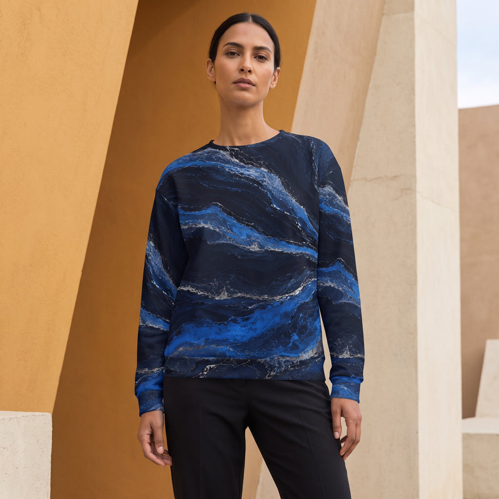A woman modeling a long-sleeve crewneck sweatshirt with a blue and navy marble wave pattern, paired with black pants, set against a modern architectural background of warm beige columns.