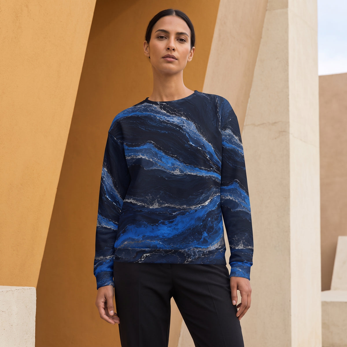 A woman modeling a long-sleeve crewneck sweatshirt with a blue and navy marble wave pattern, paired with black pants, set against a modern architectural background of warm beige columns.