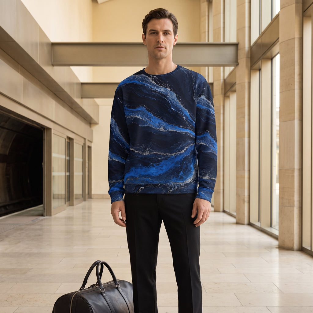 A man wearing a navy blue long-sleeve crewneck sweater with a blue and white marble wave pattern, paired with black trousers, standing in a modern stone hallway next to a black leather duffel bag.