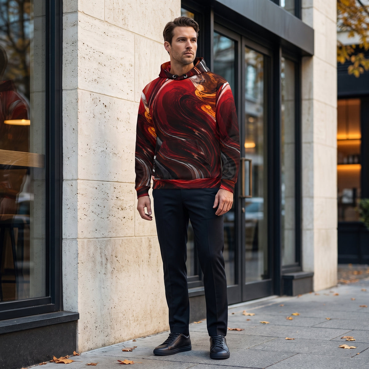 A man wearing a red and black abstract swirl-patterned hoodie, black trousers, and black shoes stands on a city sidewalk in front of a modern building with stone walls and glass doors.