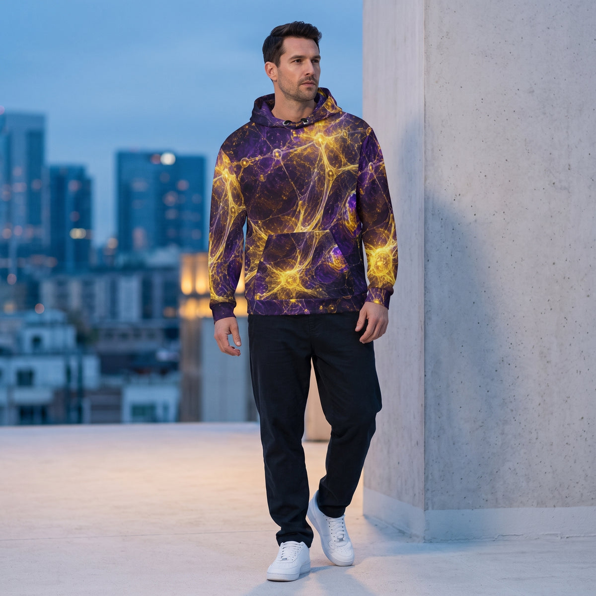 A man wearing a purple and yellow abstract patterned hoodie, black sweatpants, and white sneakers stands on a rooftop terrace with a blurred city skyline in the background.