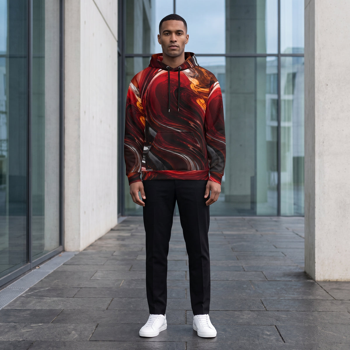A man wearing a red and black abstract swirl patterned hoodie, black pants, and white sneakers stands on a paved walkway in front of a modern glass building.
