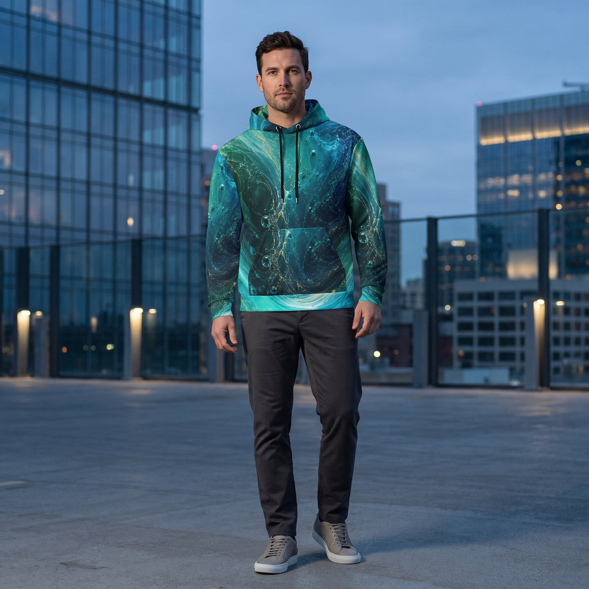 A man stands on a rooftop wearing a teal and green marbled hoodie, dark gray pants, and gray sneakers, with a city skyline in the background.