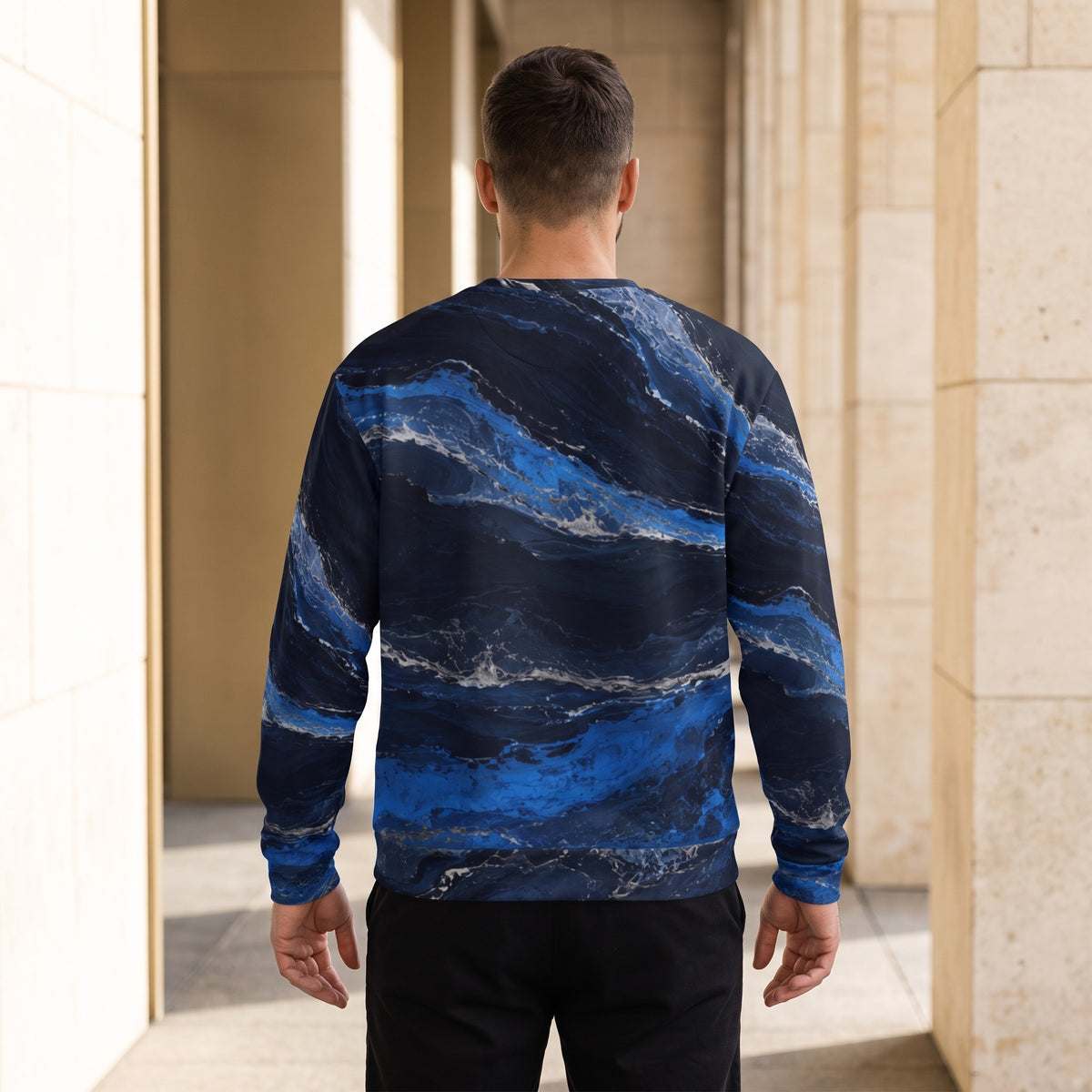 A man viewed from behind wearing a long-sleeve crewneck sweatshirt with a blue and navy marble pattern, standing in a modern architectural corridor with beige stone columns.