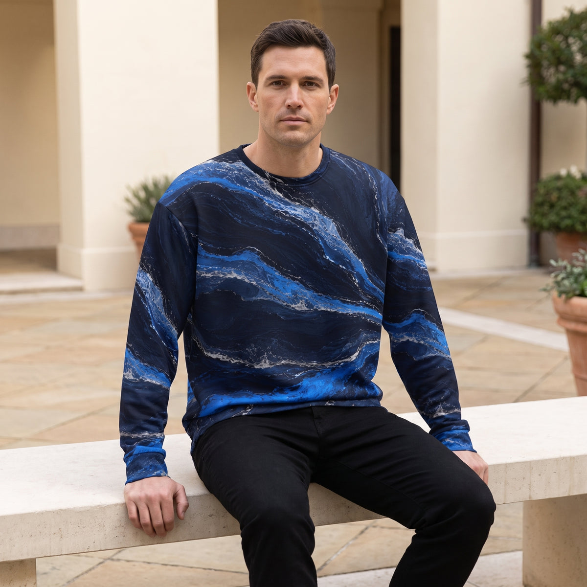 A man sitting on a stone bench wearing a long-sleeve crewneck sweatshirt with a blue and navy marble pattern, paired with black pants, in an outdoor courtyard setting.