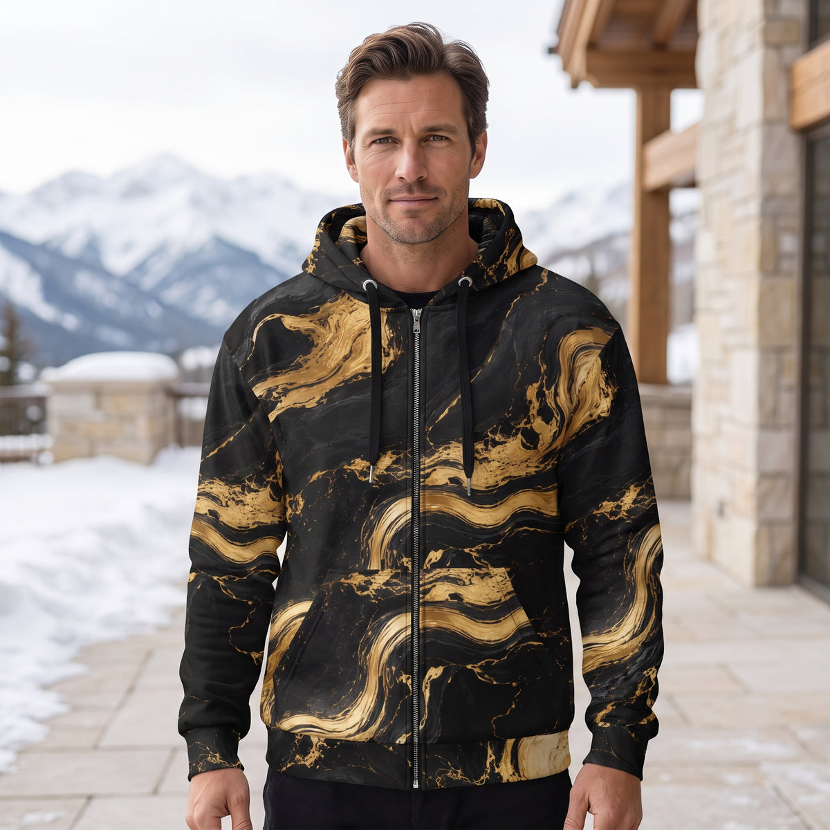 A man wearing a black and gold marble-patterned zip-up hoodie stands outdoors in front of a stone building and snowy mountains.