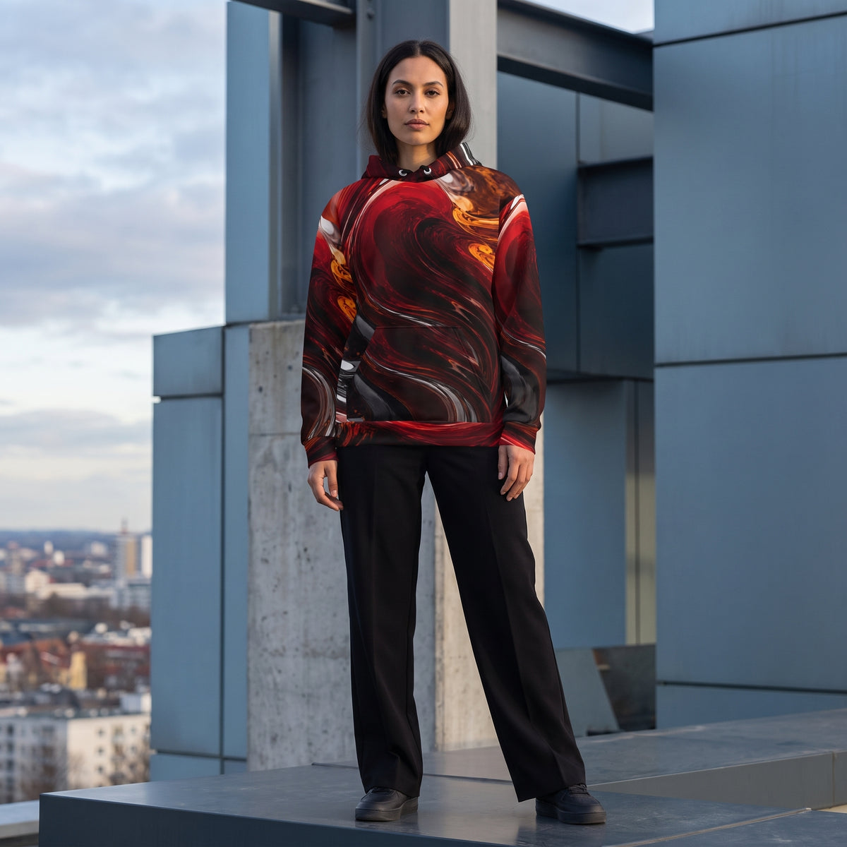 A woman stands on a rooftop wearing a pullover hoodie with a bold red, black, and orange abstract swirl pattern, paired with black wide-leg trousers and black shoes.