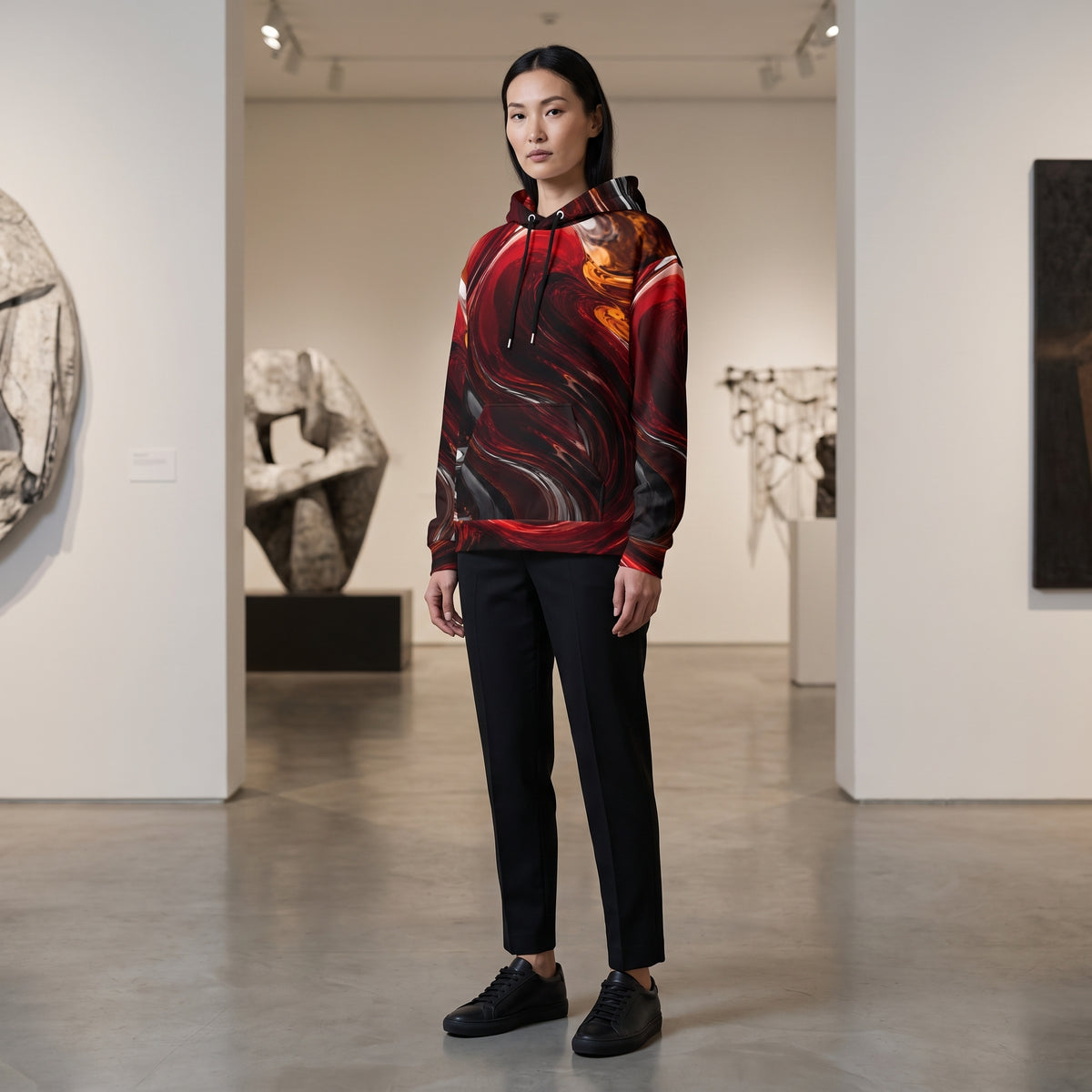 A model stands in a modern art gallery wearing a black hoodie with a red, orange, and black abstract swirl pattern, paired with black trousers and black sneakers.