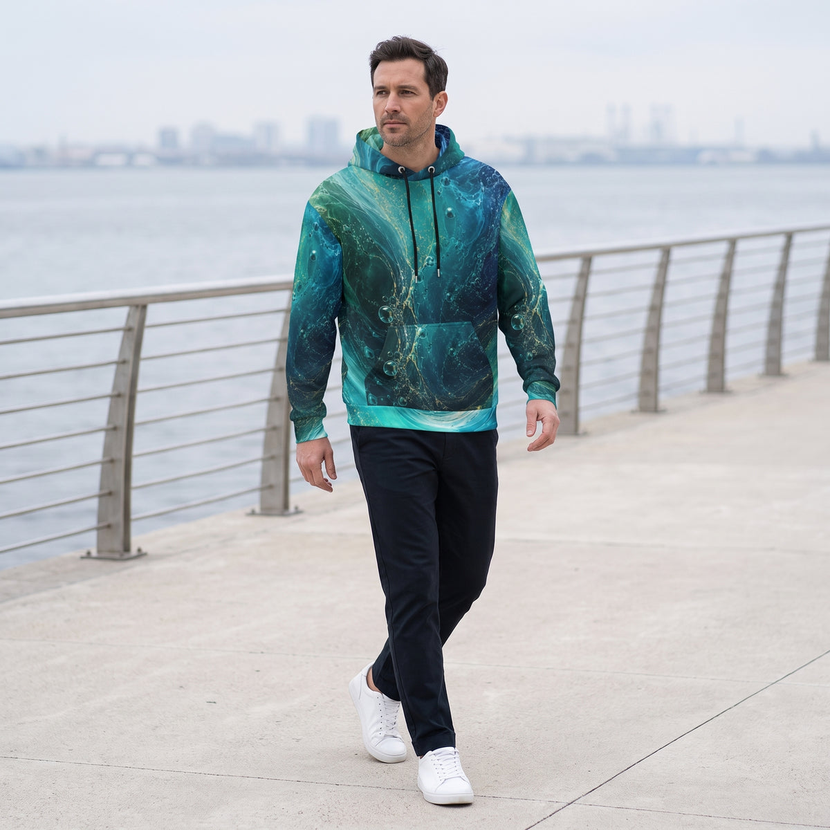 A man walking on a waterfront promenade wearing a teal and green galaxy-print hoodie, dark pants, and white sneakers.
