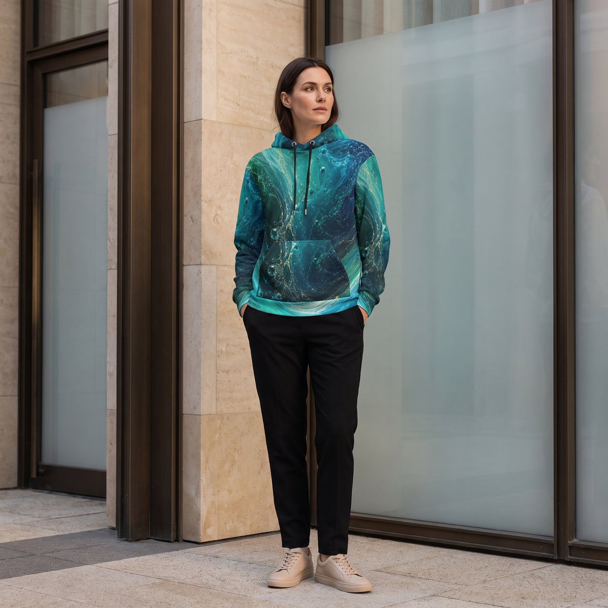 A woman stands outdoors in front of a modern glass and stone building, wearing a teal and blue abstract-patterned pullover hoodie, black pants, and beige sneakers.