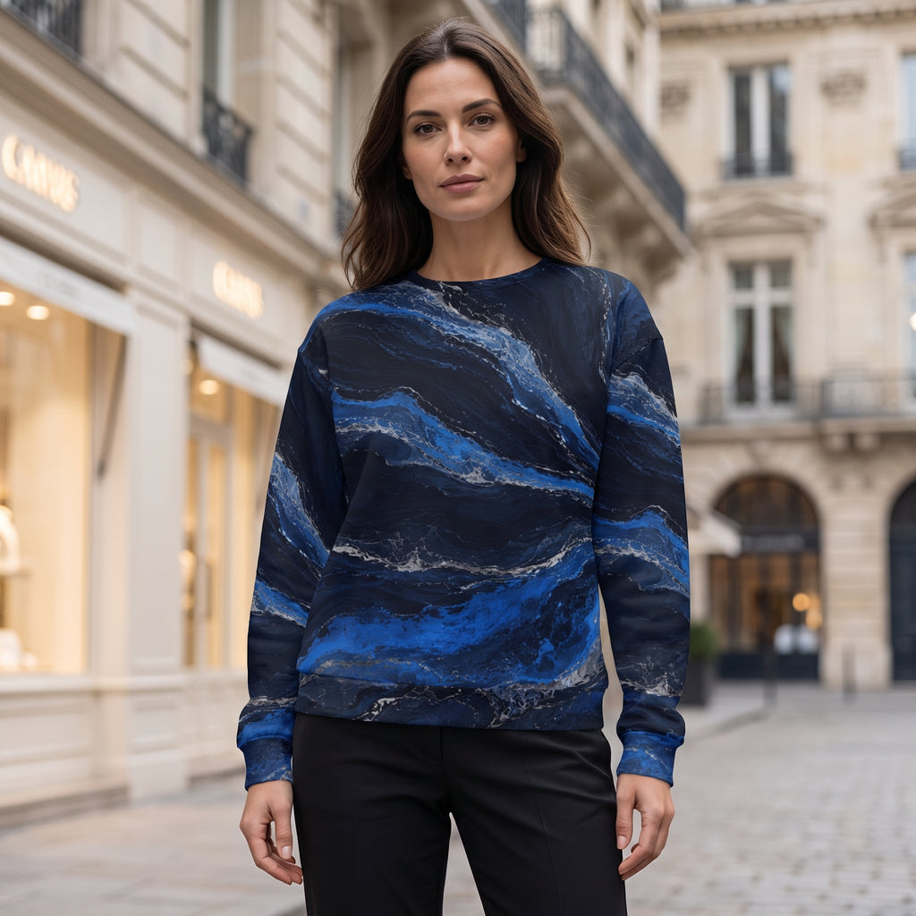 A woman stands on a city street wearing a long-sleeve crewneck sweatshirt with a blue and navy abstract marble pattern, paired with black pants.