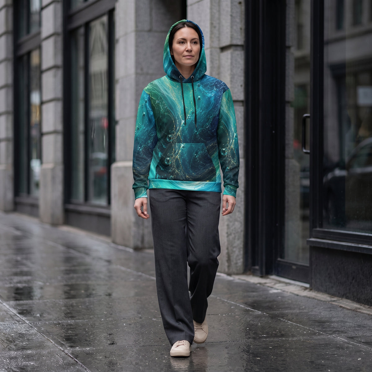 A woman walking on a wet city sidewalk wearing a teal and green marble-patterned hoodie with dark gray pants and beige sneakers.