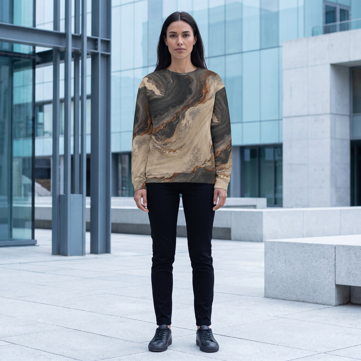 A woman stands outdoors in front of a modern glass building, wearing a long-sleeve crewneck sweatshirt with a brown and beige marble pattern, black pants, and black shoes.