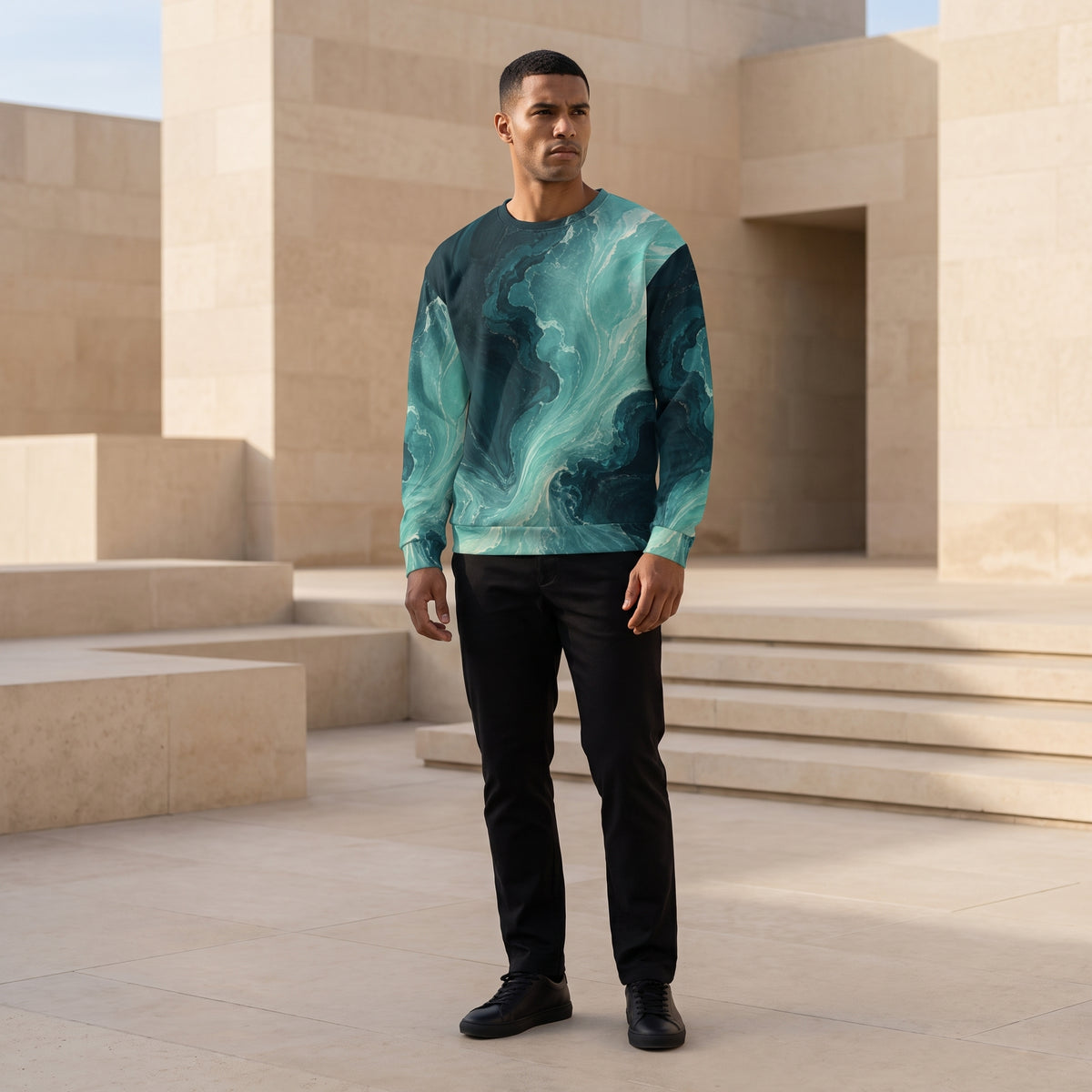 A man wearing a long-sleeve crewneck sweatshirt with a teal and dark green marble swirl pattern, paired with black pants and black shoes, standing in a modern architectural setting with beige stone steps.