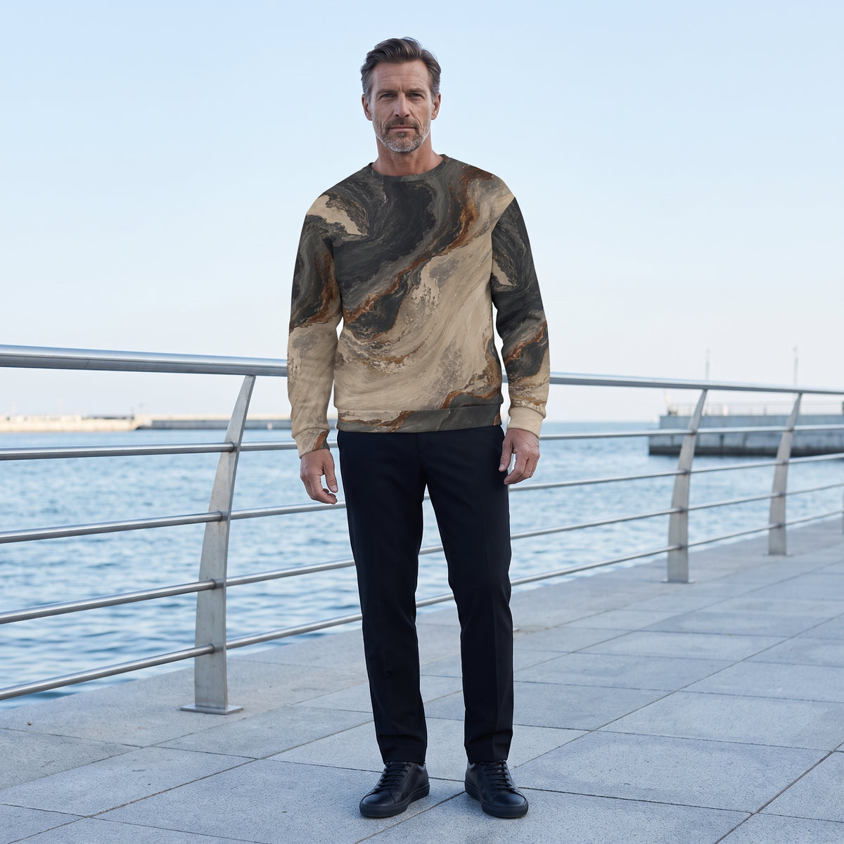 A man stands on a waterfront promenade wearing a long-sleeve crewneck sweatshirt with a brown and beige marbled pattern, paired with black trousers and black shoes.