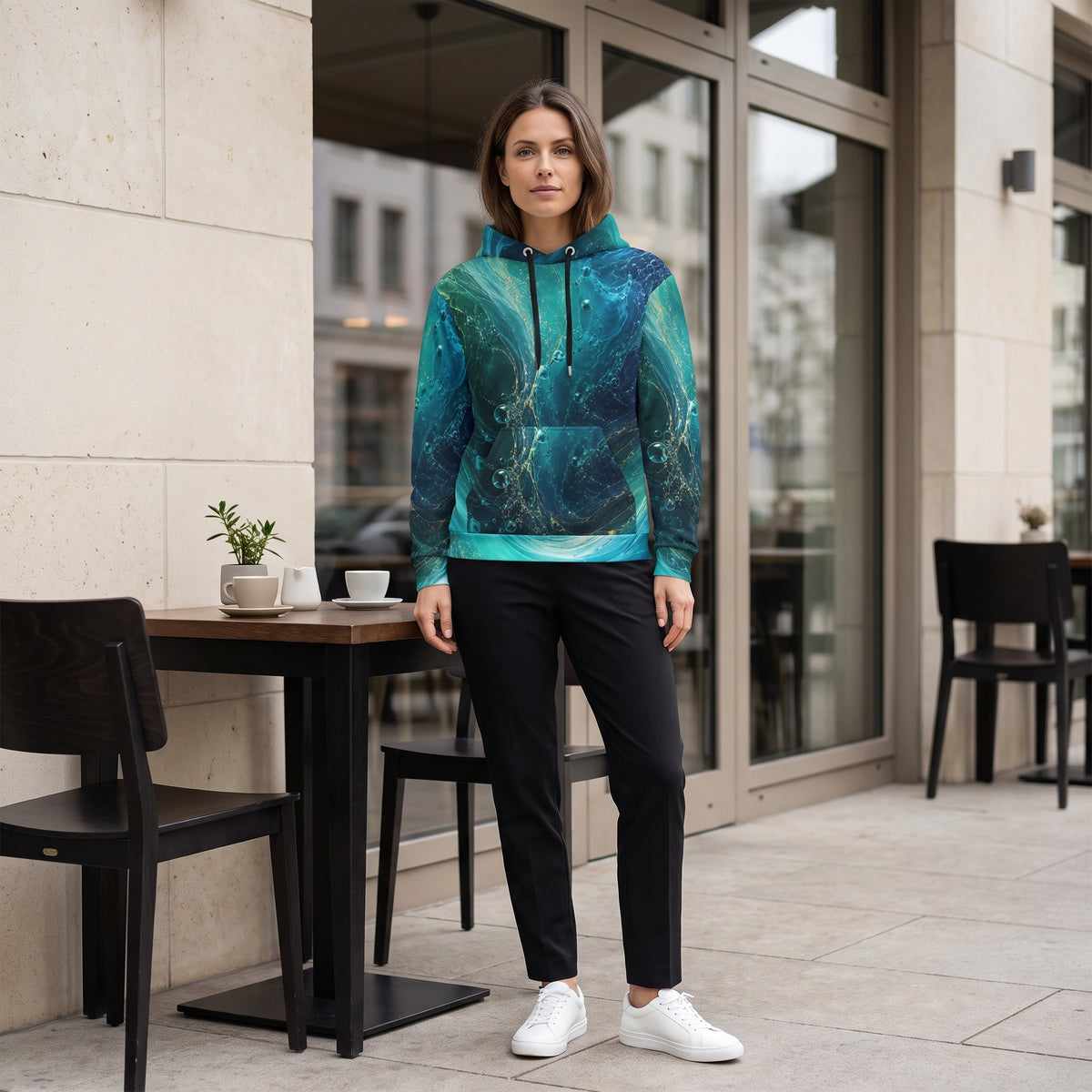 A woman stands on a patio outside a modern building, wearing a teal and blue abstract-patterned hoodie, black pants, and white sneakers.