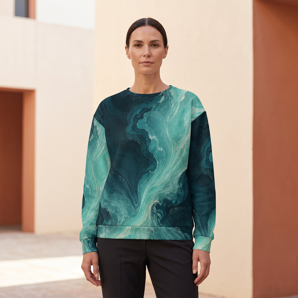 A woman modeling a long-sleeve crewneck sweatshirt with a teal and dark green marble swirl pattern, paired with black pants, set against a modern beige and terracotta building background.