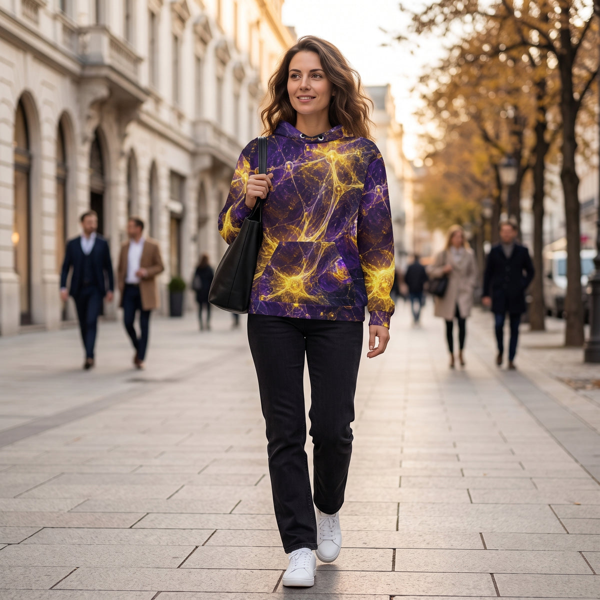 A woman walking down a city street wearing a purple and yellow abstract patterned hoodie, black pants, and white sneakers, carrying a black shoulder bag.