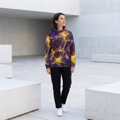 A woman stands in a modern concrete courtyard wearing a purple and yellow abstract patterned hoodie, black pants, and white sneakers.
