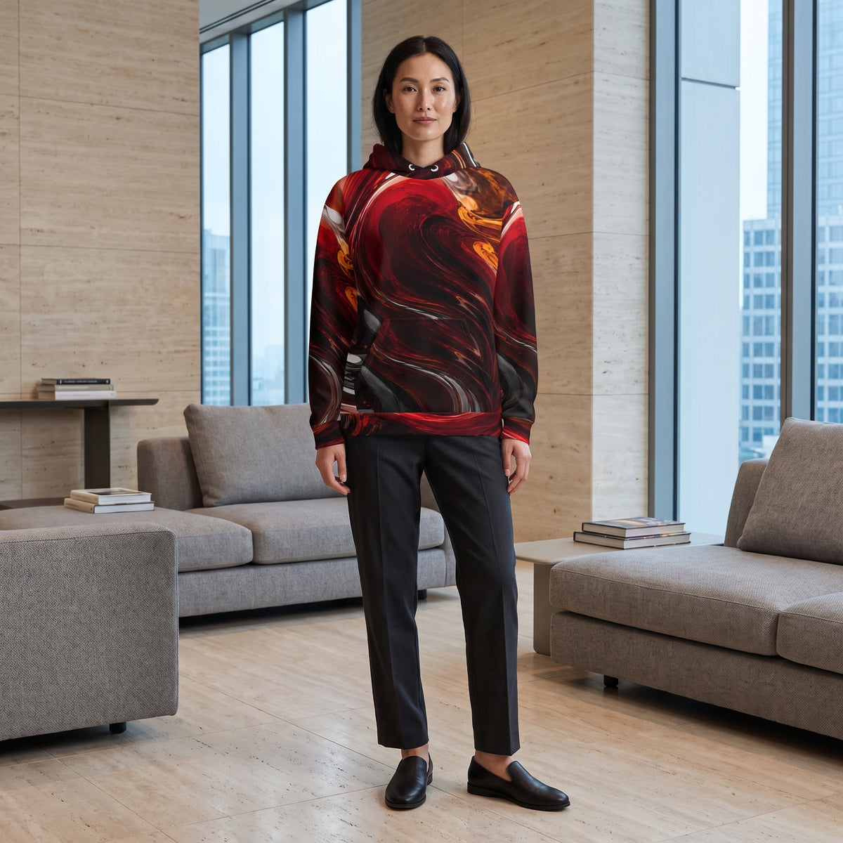 A woman stands in a modern living room wearing a red and black abstract-patterned hoodie, dark charcoal trousers, and black loafers.