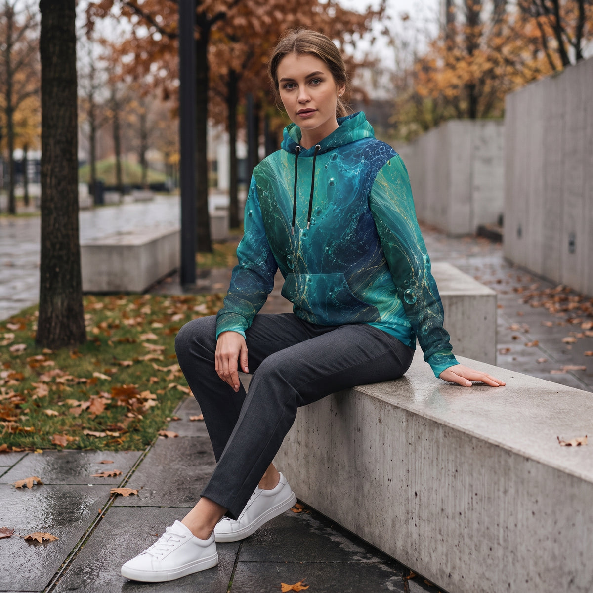 A woman sits on a concrete bench in an outdoor park setting, wearing a teal and blue marbled hoodie, dark gray pants, and white sneakers.